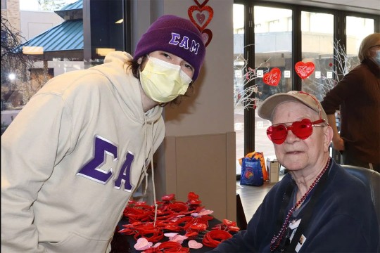 a student wearing a sweatshirt with the Sigma Alpha Mu Delta Omega chapter logo poses with a senior citizen at St Johns Home for a Valentines Day celebration