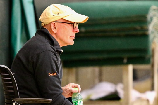 A side profile of adjunct professor Ed Sherman. He's wearing a white baseball hat, black glasses, and a black zip-up sweater with the word coach embroidered in orange on the bicep.