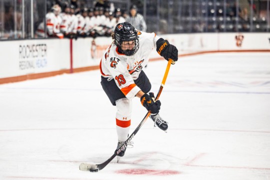 Ice hockey player in a white and orange RIT jersey skates forward, controlling the puck with a black and yellow hockey stick, while teammates watch from the bench in the background.