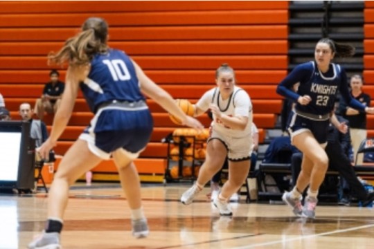 three college age girls play basketball on a brightly lit, collegiate court. One wears white with orange, the other two wear navy blue with white.