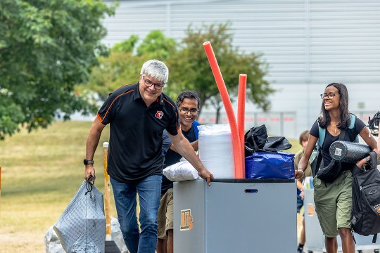 Smiling people push a cart filled with dorm supplies outside an R I T building.