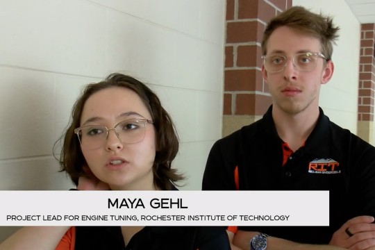 two students in R I T shirts stand next to a white wall.