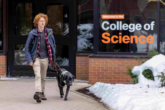 a man with red hair walks with a black dog outside a building labeled College of Science