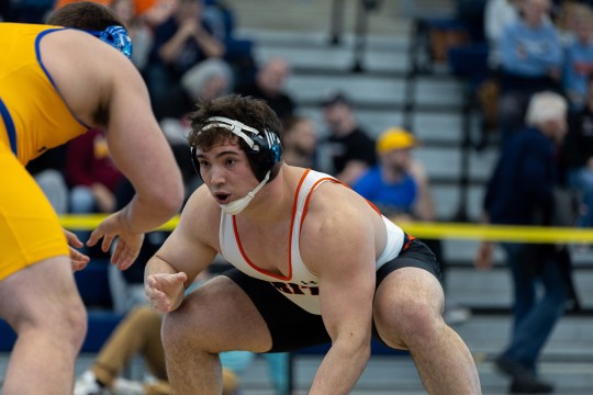 a college age male in a white and orange wrestling uniform circles and spars with another wrestler wearing yellow.