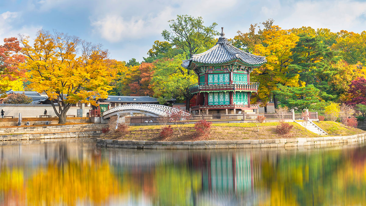 Traditional Korean pavilion by a calm pond, surrounded by vibrant autumn trees and colorful reflections in water.