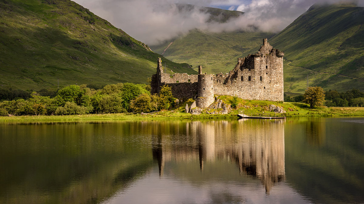 Ancient stone castle reflected in a still lake, surrounded by lush green hills and misty mountain peaks.