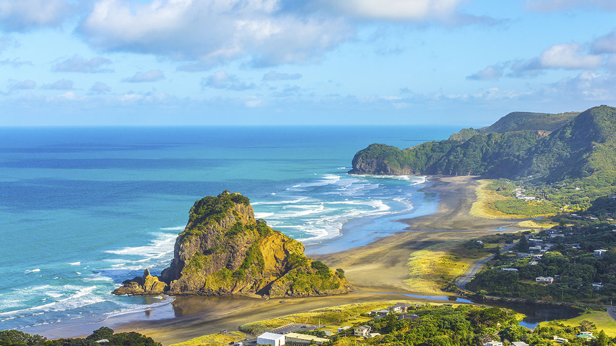 Lion Rock on Piha Beach with crashing waves, lush green hills, and scattered homes along New Zealand’s coastline.