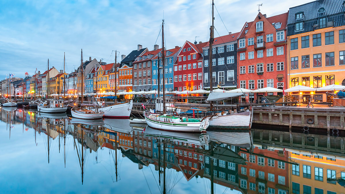 Colorful buildings and moored sailboats reflect on the calm waters of Nyhavn Harbor in Copenhagen at dusk.