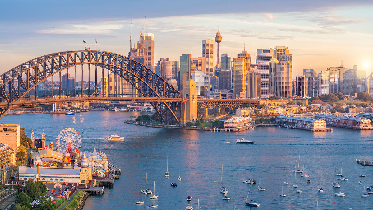 Sydney Harbour at sunset with boats, the Harbour Bridge, city skyline, and Luna Park in view.