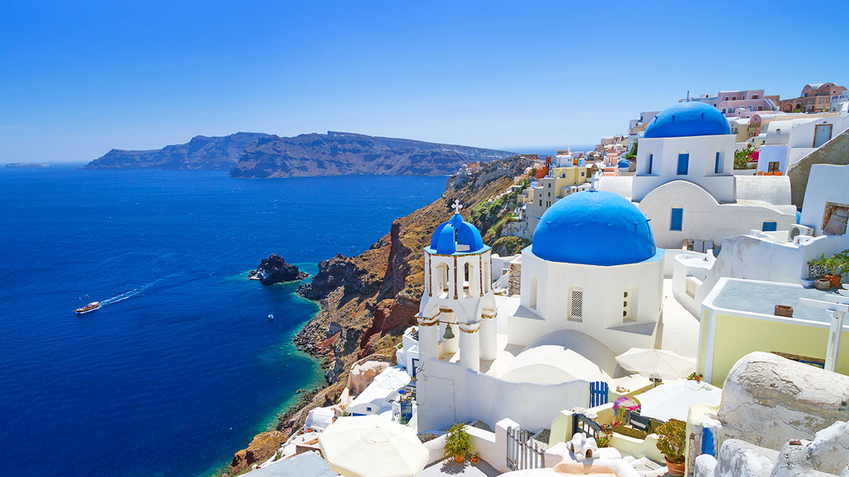 Whitewashed buildings with blue domes overlook the deep blue Aegean Sea on a sunny day in Santorini.