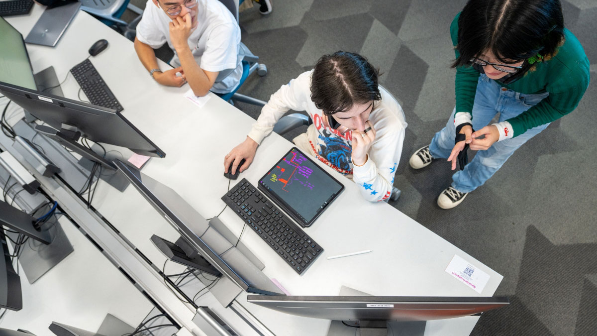 Three students collaborating on a coding project in a computer lab, with screens displaying programming work.