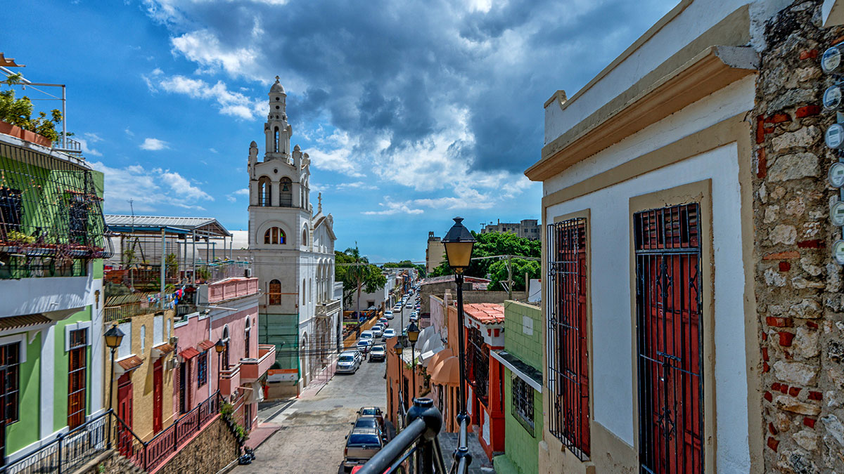 Colorful colonial buildings and a white church tower lining a vibrant street under a partly cloudy sky.