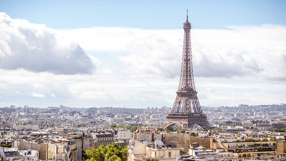 The Eiffel Tower rising above the Parisian skyline under a bright blue sky with scattered clouds.