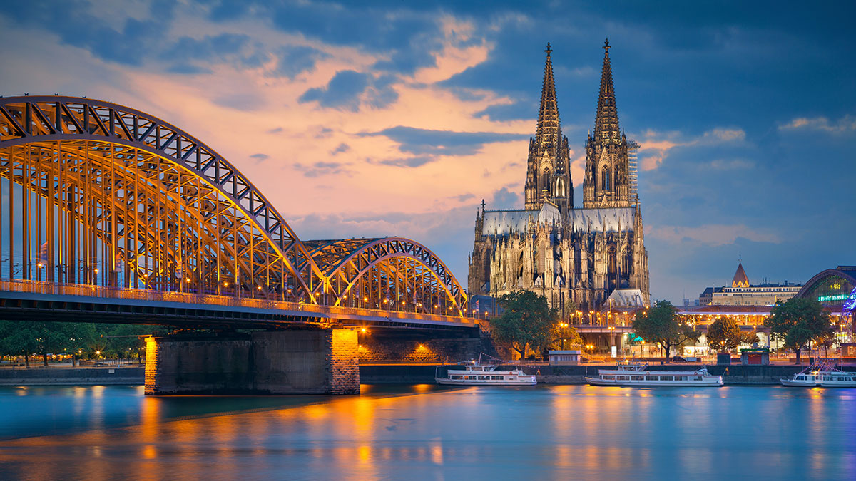 Cologne Cathedral and the Hohenzollern Bridge illuminated at sunset, reflecting on the Rhine River.