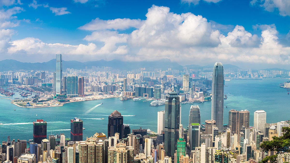 A panoramic view of Hong Kong’s skyline, Victoria Harbour, and distant mountains under a bright, partly cloudy sky.