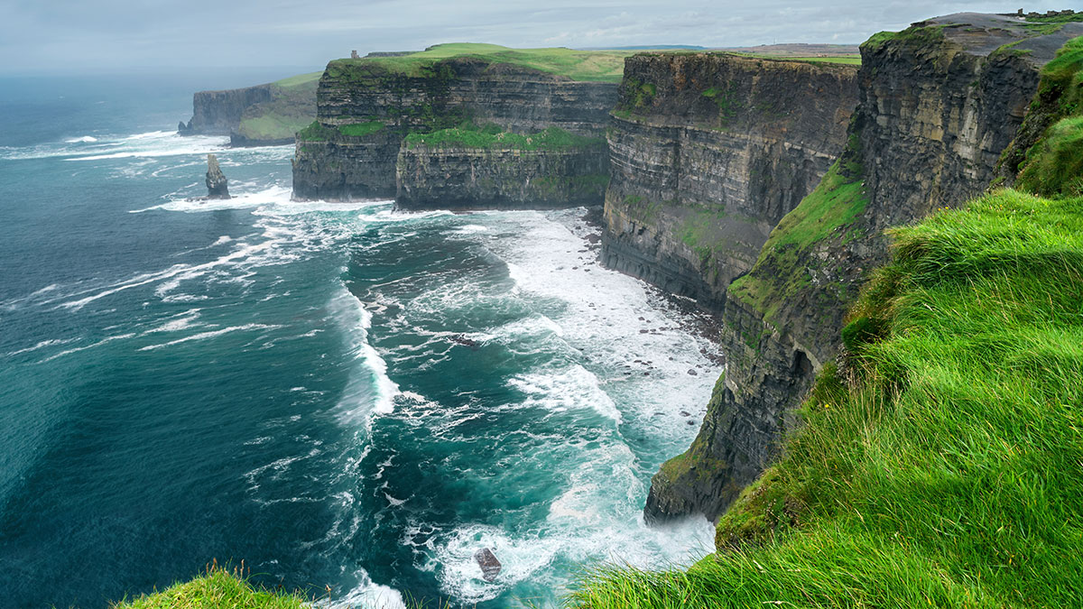 The dramatic Cliffs of Moher in Ireland, with lush green grass and waves crashing against the rocky shoreline.