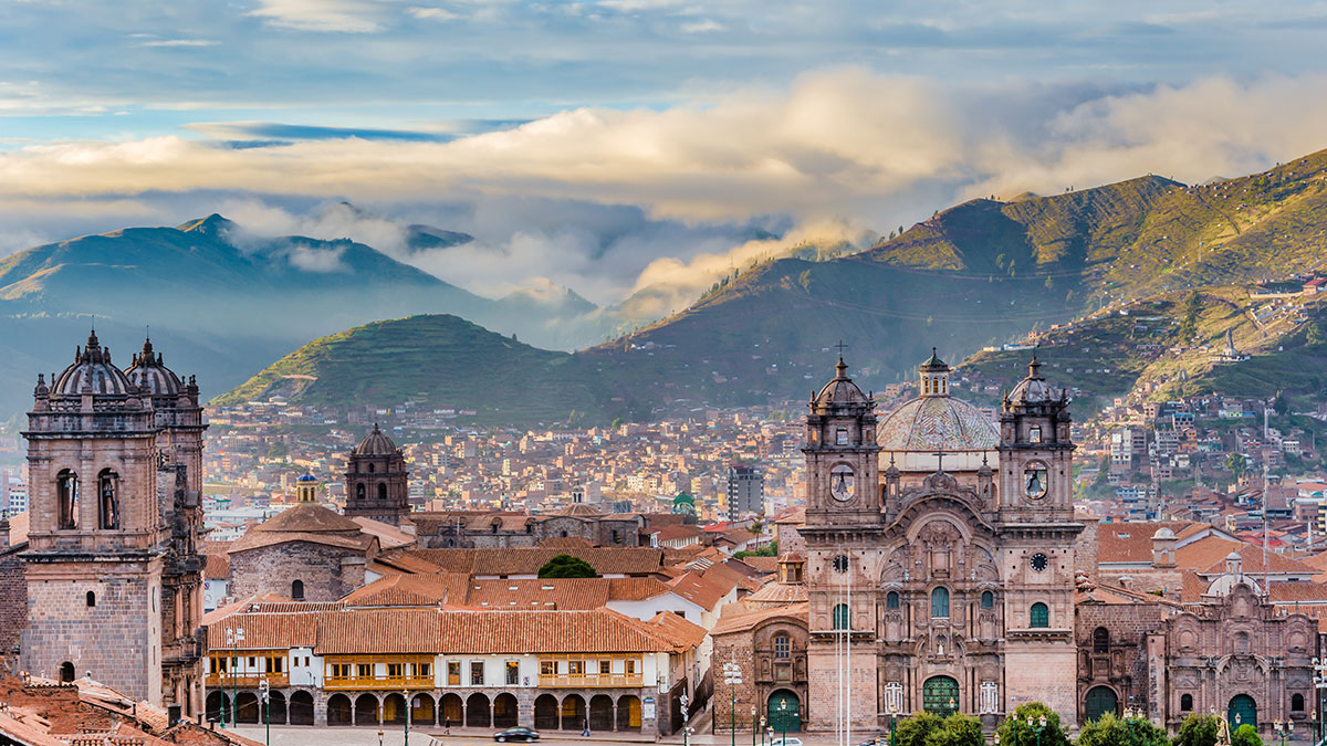 Cusco's historic Plaza de Armas, featuring colonial churches, red-tiled roofs, and misty Andean mountains in the background.