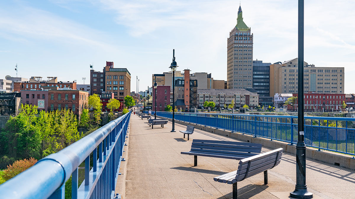 Pedestrian bridge in Rochester, New York, featuring benches, lampposts, and a city skyline with historic buildings under a clear sky.