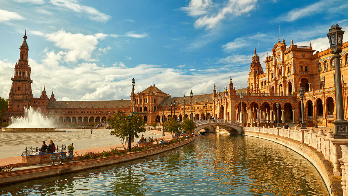 Plaza de España in Seville, Spain, featuring a grand architectural facade, fountain, canal, and ornate bridges under a bright sky.