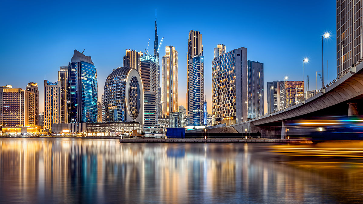 Dubai skyline at dusk, featuring modern skyscrapers, illuminated reflections on calm waters, and a bridge with passing traffic lights.