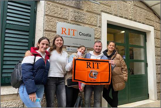 Five students pose outside the RIT Croatia campus holding an orange RIT flag, with a stone building backdrop.