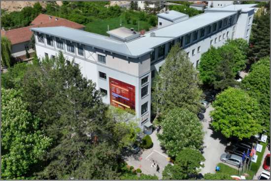 An aerial view of the RIT Kosovo campus, featuring a modern gray building surrounded by lush green trees and pathways.