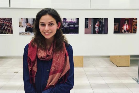 Lauren Peace poses for a photo in the University Gallery. She stands in front of a board with five photos lined up next to each other.