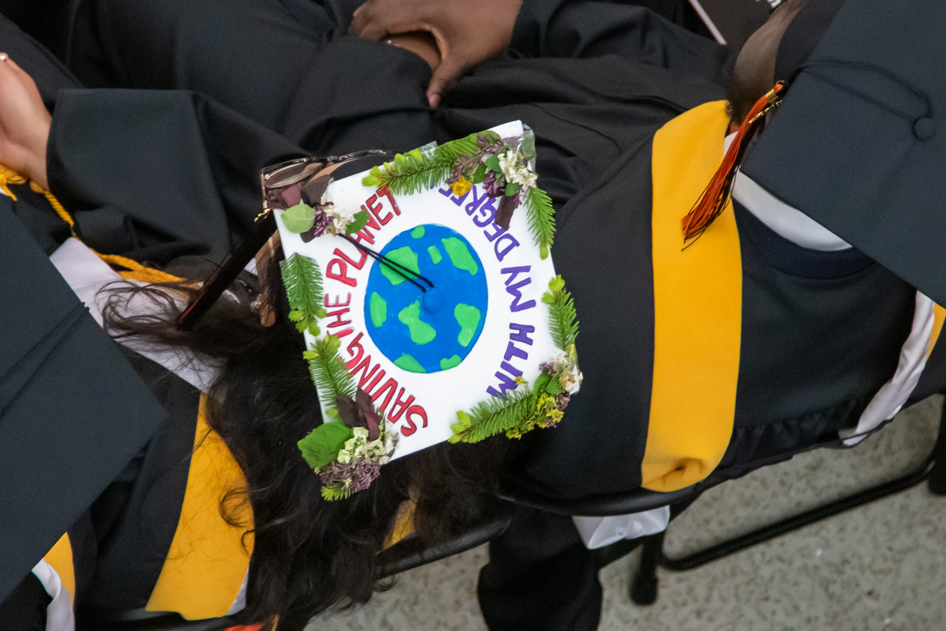 Aerial close up of the top of a graduating student's grad cap that has a painting of a globe with the words 'saving the planet with my degree'