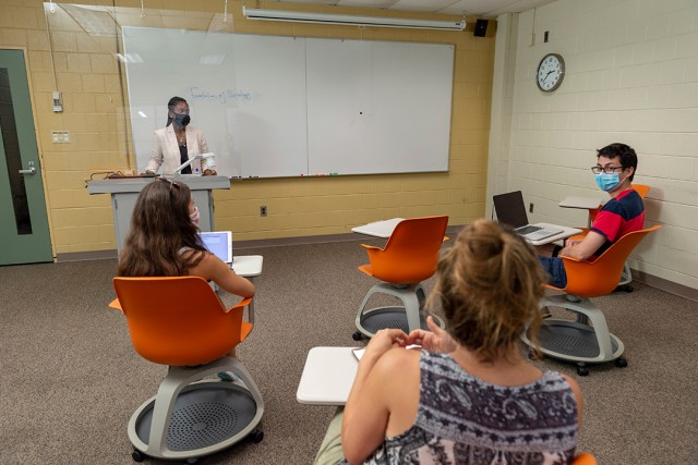 professor wearing a mask stands in the front of a classroom behind a plexiglas barrier as three students sit six feet apart.