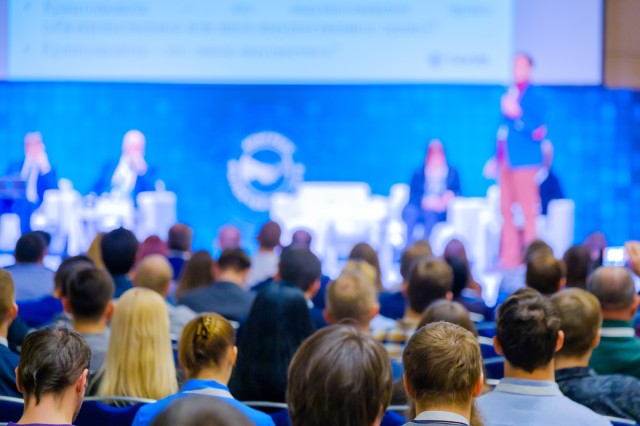 View of conference panel discussion from afar with rows of seated attendees seen from behiind