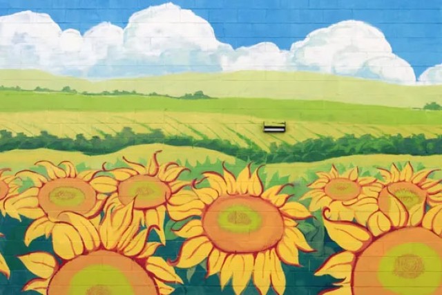 a brick wall covered by a mural of a green field in the background and sunflowers in the foreground.