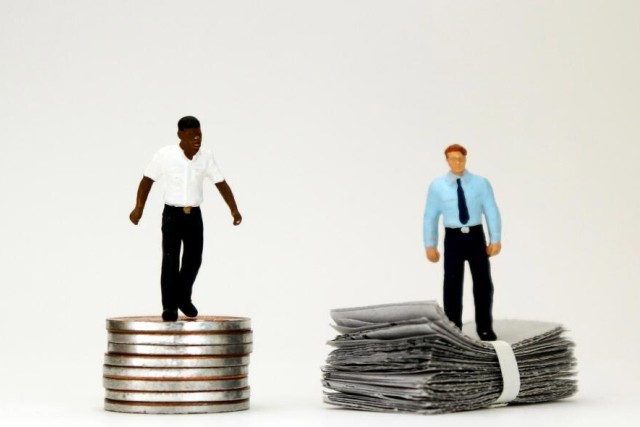 on the left, a plastic figuring of a black man stands on a pile of coins. On the right, a plastic figuring of a white man stands on a stack of paper money.