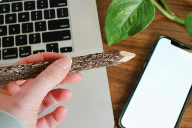 A photo image shows a hand holding a large, rough-hewn pencil over a laptop keyboard, with a mobile phone next to it.