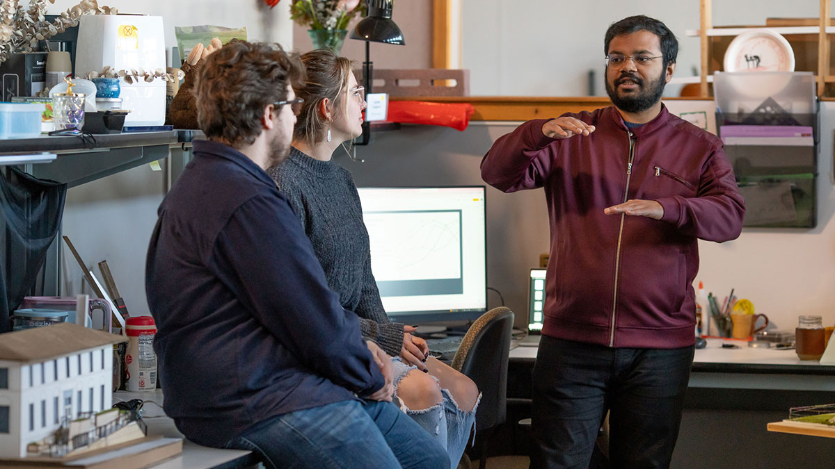 A student gestures while speaking to two other students in a modern workspace with a computer in the background.