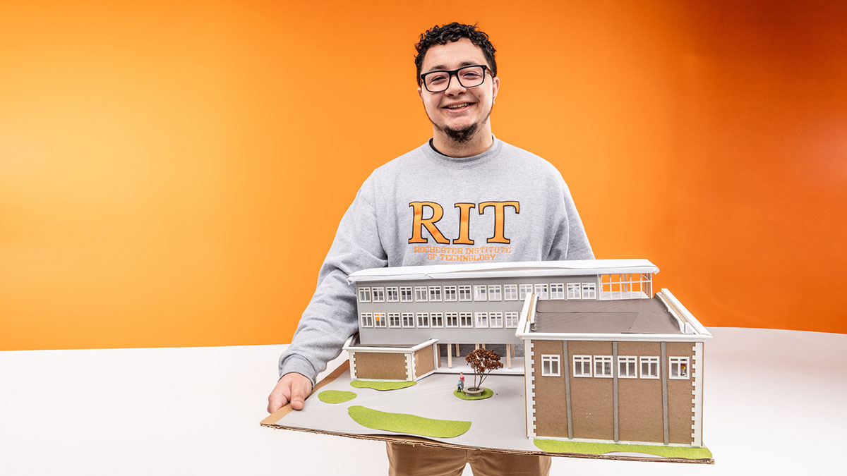 A smiling student wearing an RIT sweatshirt holds a detailed architectural model in front of an orange background.