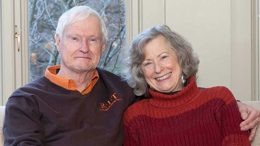 An older couple, both smiling, sit closely together in a well-lit room with large windows behind them.