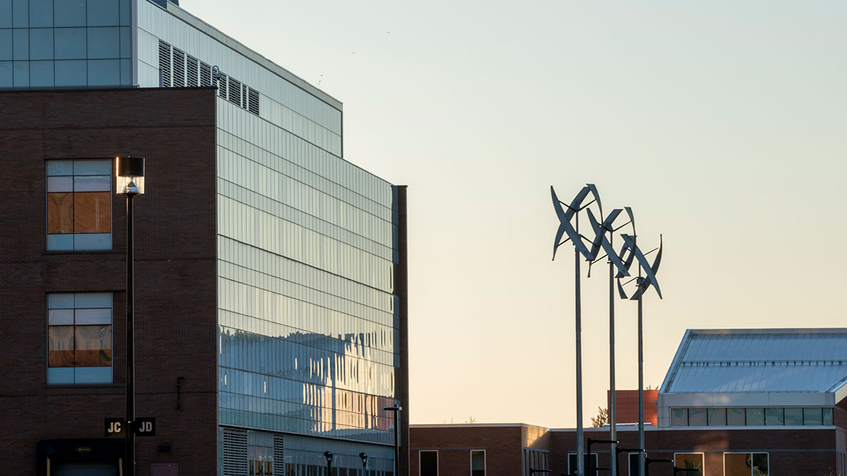 A modern glass building with three vertical wind turbines beside it, photographed during sunset.