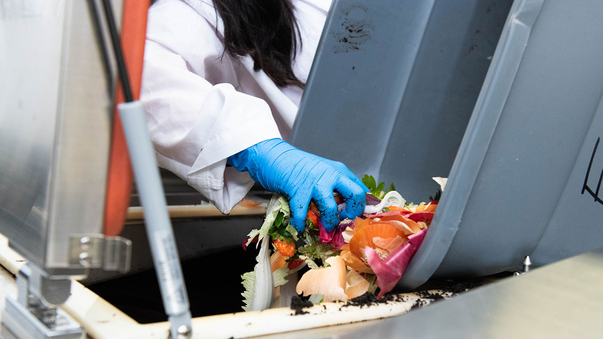 A gloved hand reaches into a compost bin filled with food scraps including vegetable peels.