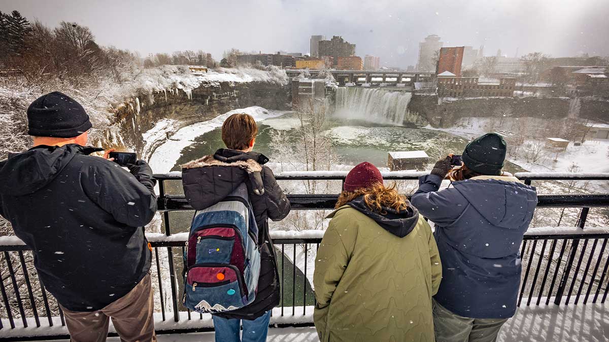 Four people stand at a snowy overlook photographing a large waterfall with a city skyline in the background.
