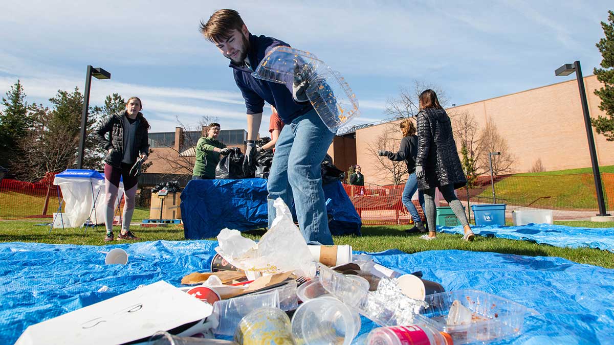a student throws a take-out container into a pile of trash as a team of volunteers sorts through recyclables.