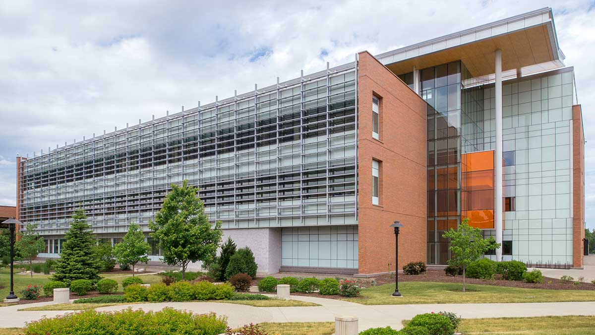 A modern brick and glass building is seen on a bright day, surrounded by manicured trees and pathways.