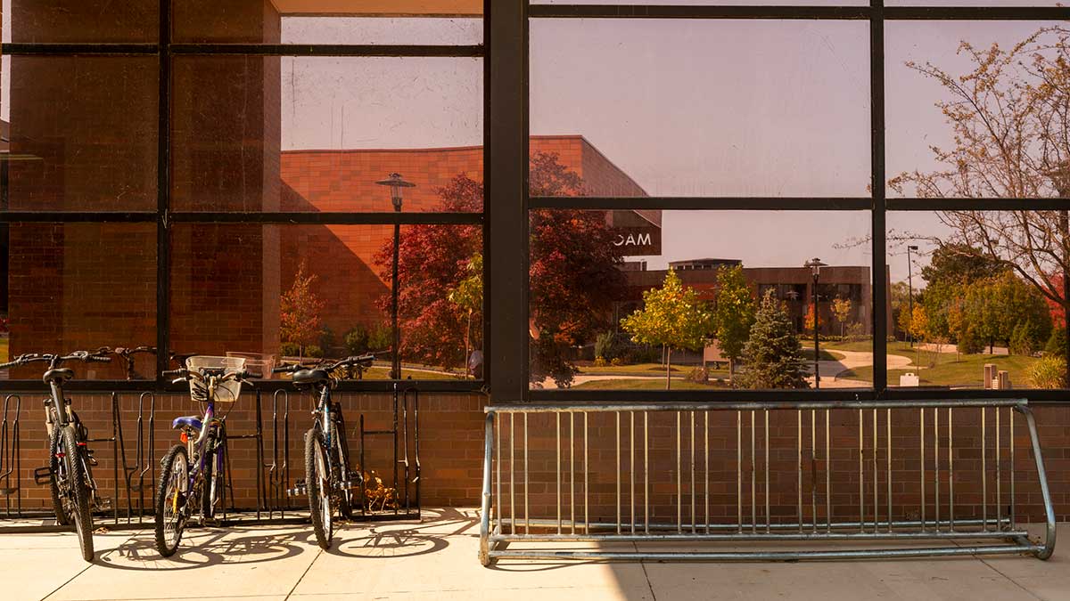 Several bicycles are parked in a rack in front of large windows reflecting a landscaped campus scene.