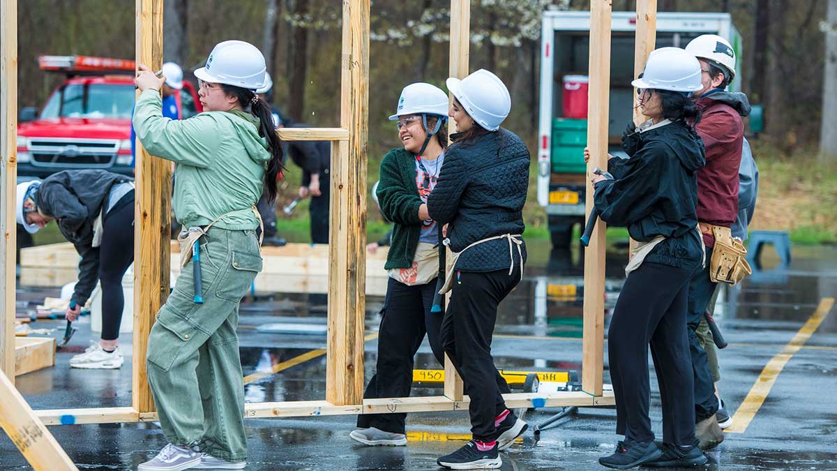 student volunteers wearing hard hats carry a framed wall for a Habitat for Humanity project.