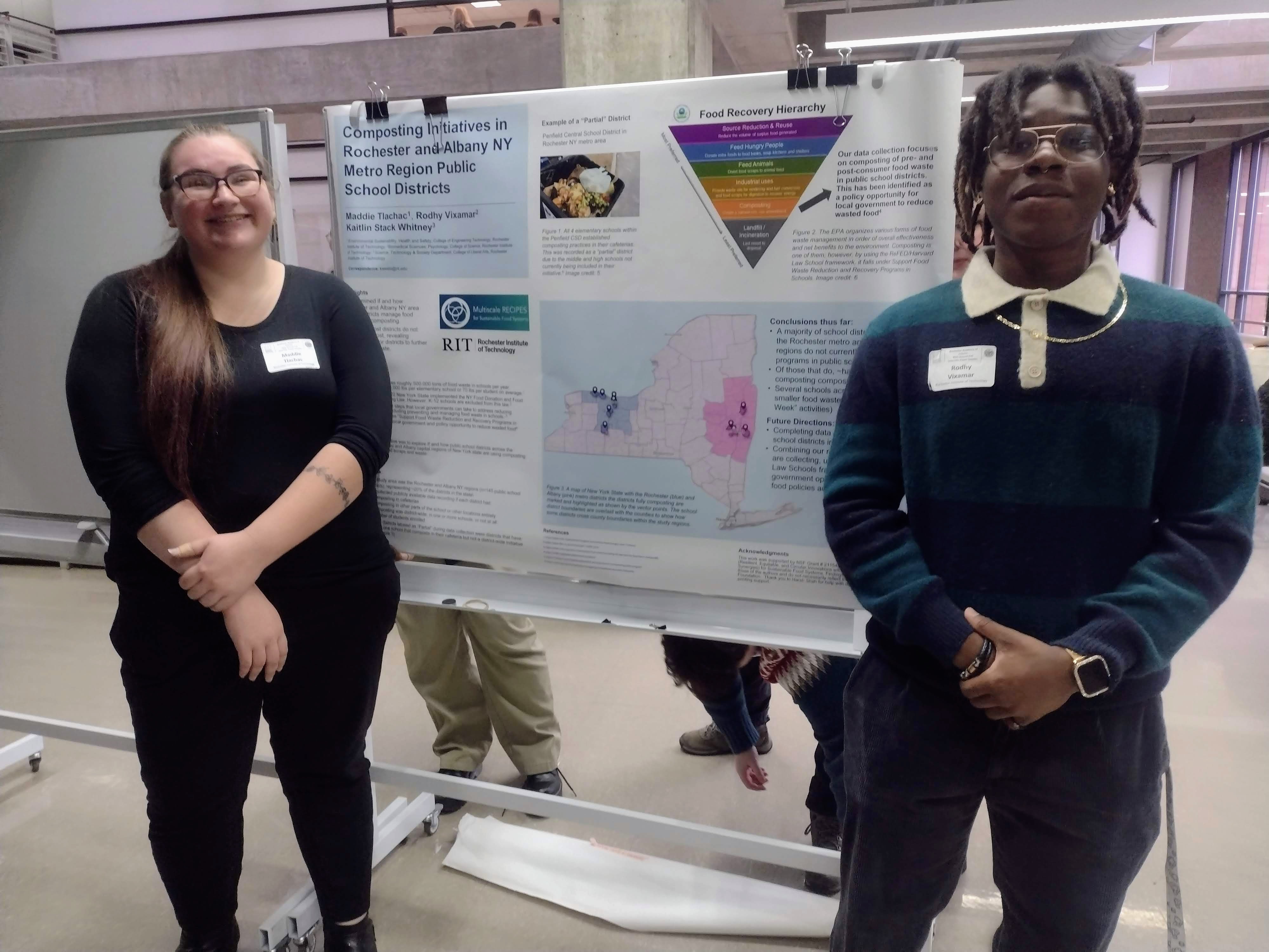 two people in dark clothes and glasses (Maddie and Rodhy) smile at the camera, standing with their research poster between them