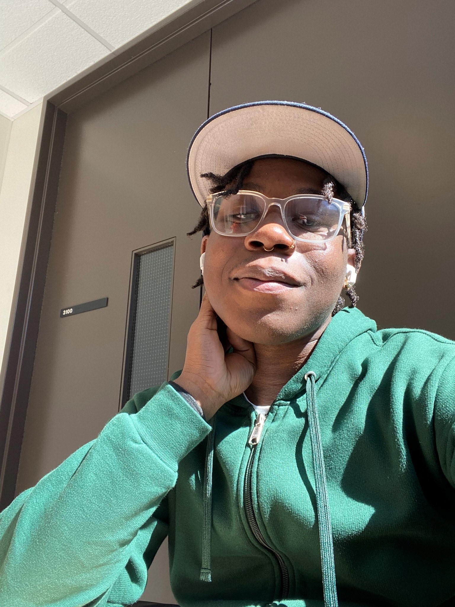 a smiling Black man with nose rings and glasses in a baseball cap and green sweatshirt