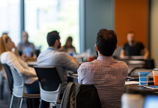 A group of adults seated around a conference table in a meeting or workshop, listening to a speaker at the front of the room.