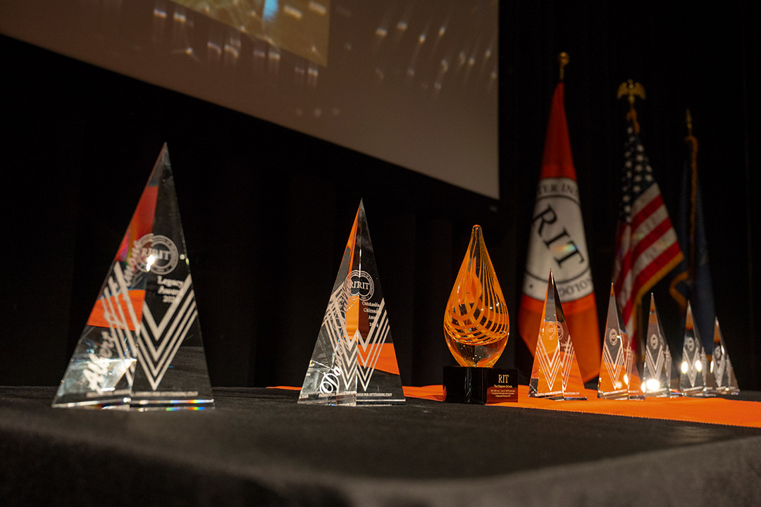 Crystal and glass awards displayed on a black table with RIT and American flags in the background.