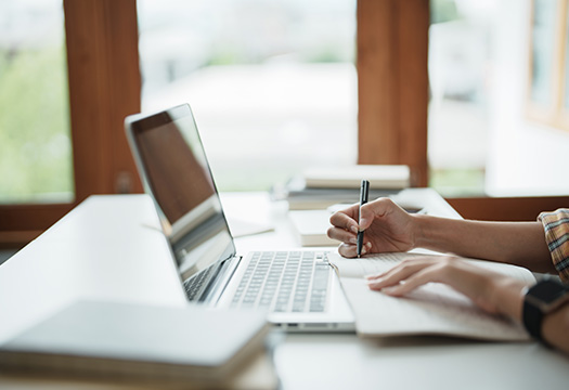Person at table, only their hands and forearms are can be seen, holding a pencil over a notebook and an open laptop. 