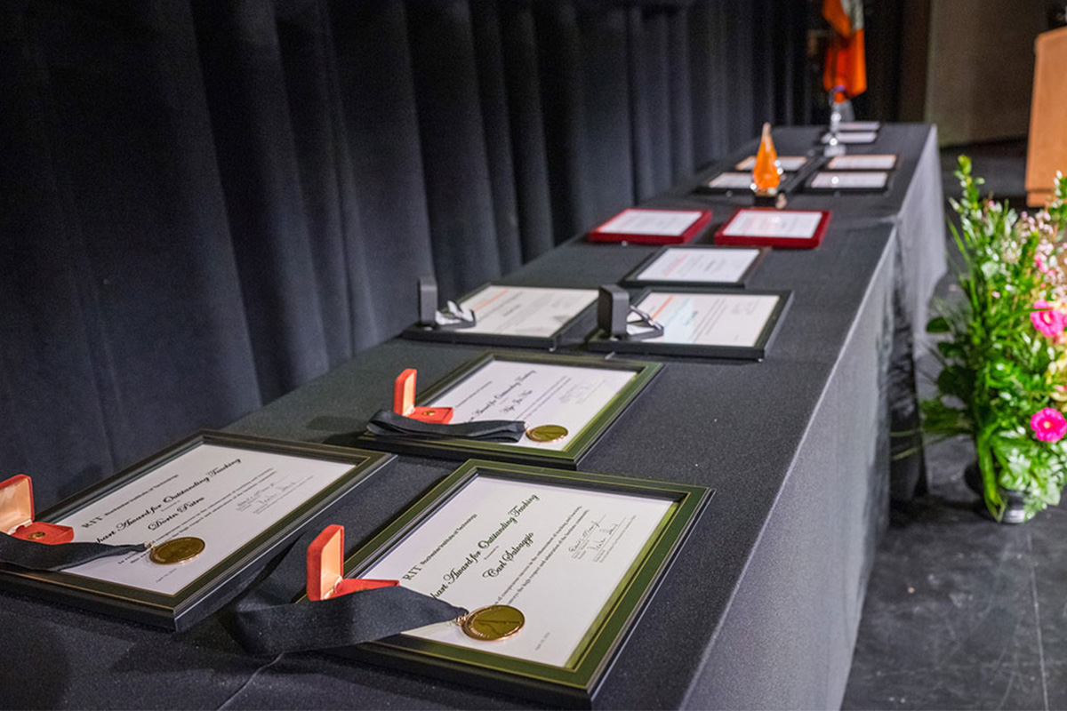 A row of certificates and awards displayed on a black-draped table with flowers in the foreground.