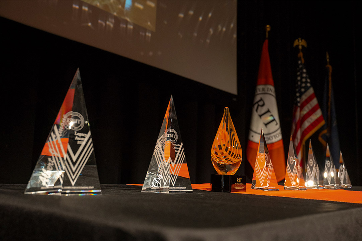 Crystal and glass awards displayed on a black table with RIT and American flags in the background.